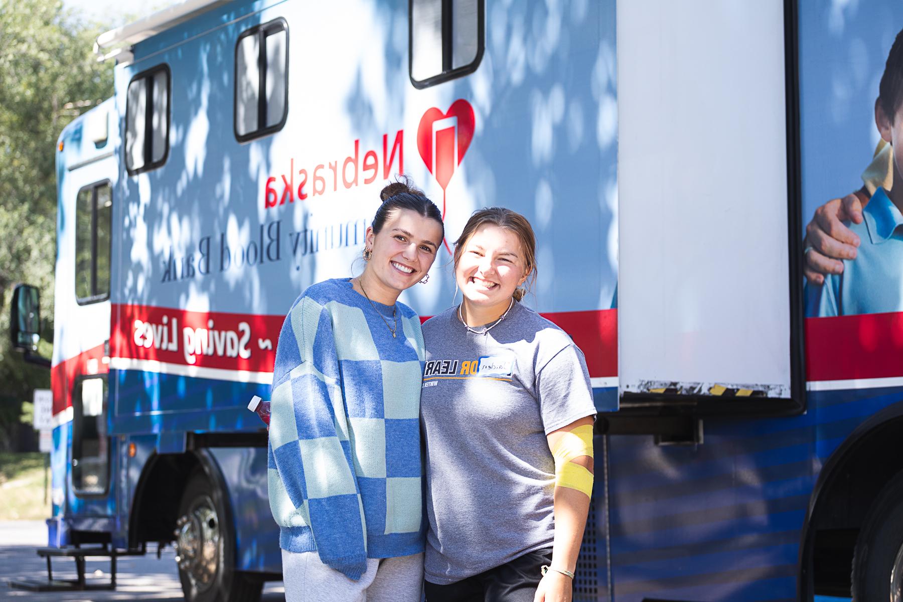 两个学生, 一个人献血后肘部缠着黄色绷带, 站在克里特岛佩里校园中心外的内布拉斯加州社区血库的血液车前. 
